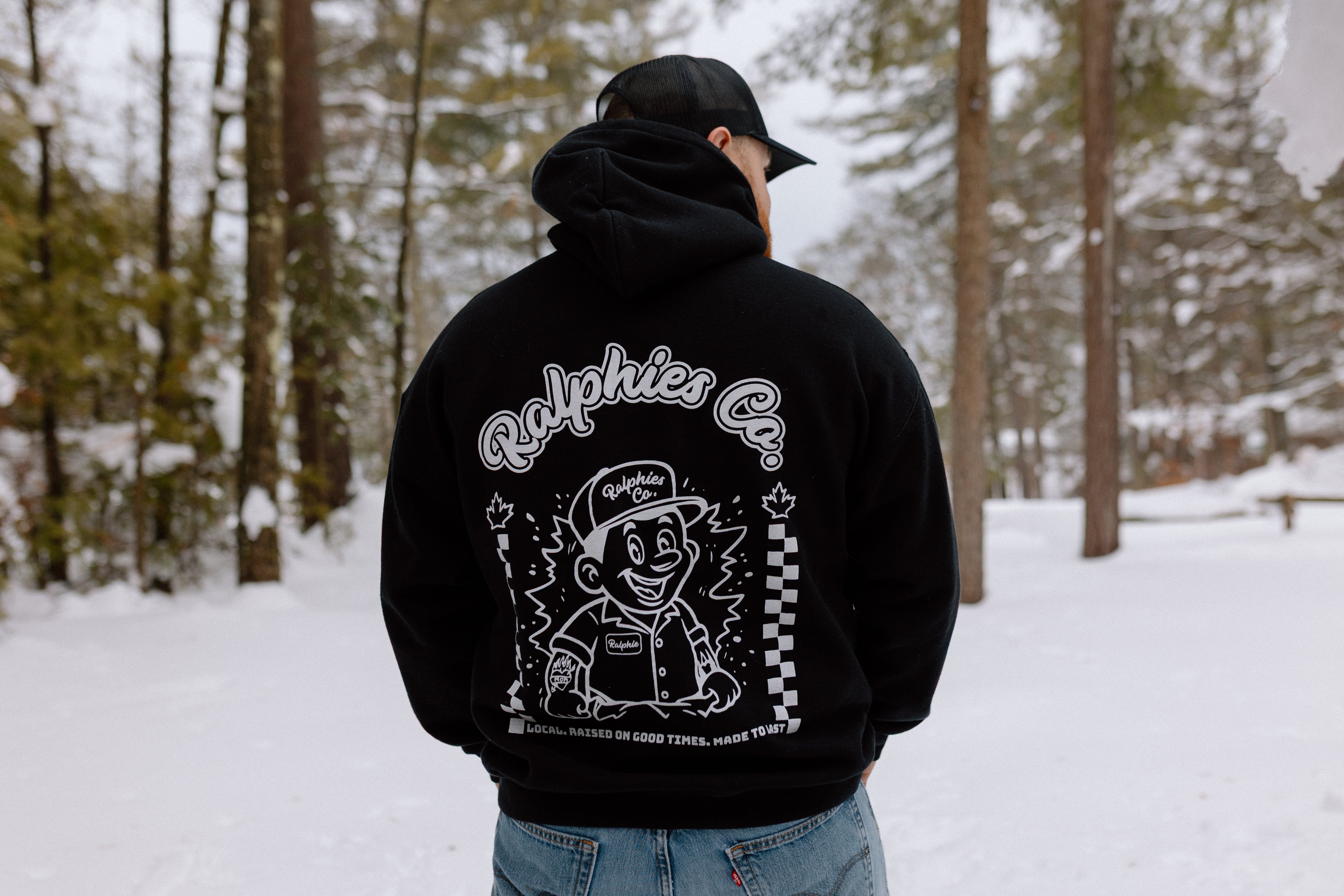 Person wearing a black Ralphies Co 
hoodie with a graphic design in a snowy forest